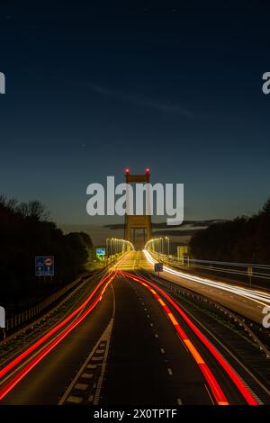 Long exposure shot of traffic light trails on a highway leading to a towering suspension bridge at dusk Stock Photo