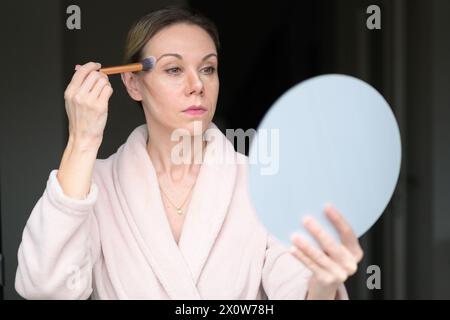 Blonde woman applying makeup with a brush and looking into a mirror at home Stock Photo