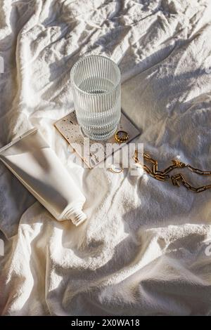 Morning routine items, glass of water, body lotion and jewelry in natural sunlight on the bed Stock Photo