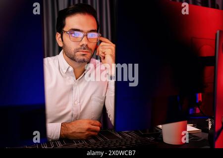 Smart businessman wearing glasses, concentrating on dynamic stock exchange investment on two pc screens at neon light office in dark room night with Stock Photo