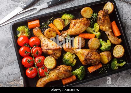 Tasty Baked ruddy chicken legs with potatoes, broccoli, tomato, carrot close-up on a baking sheet on the table. Horizontal top view from above Stock Photo