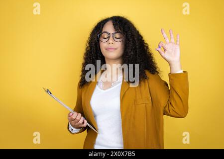 African american business woman with paperwork in hands over yellow background relax and smiling with eyes closed doing meditation gesture with finger Stock Photo