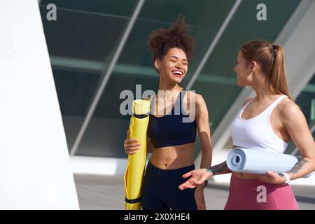 Two women in sports uniforms hold yoga mats, chatting while standing outdoors Stock Photo