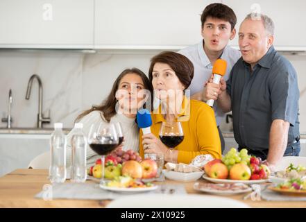 Happy family singing karaoke using microphone during family holiday, parents and adult children at festive table in kitchen Stock Photo