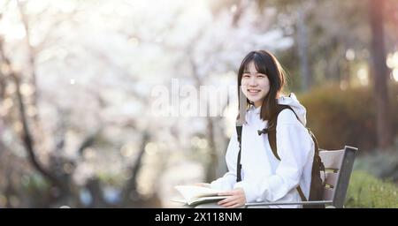 Concept of a college student sitting on a bench in a school park, reading a book and studying, and a high school student smiling while carrying a bag Stock Photo