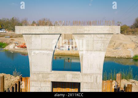 Large modern gray concrete bridge pillar in foundation surrounded by metal piles. Reinforced pole under construction is across river. Team of people w Stock Photo