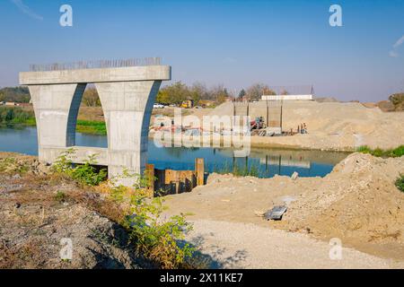 Large modern gray concrete bridge pillar in foundation surrounded by metal piles. Reinforced pole under construction is across river. Team of people w Stock Photo