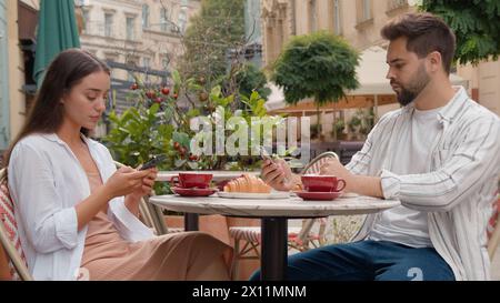 Caucasian couple in city cafe using mobile phone telephone texting working serious man woman breakfast outside together gen z drinking coffee chatting Stock Photo
