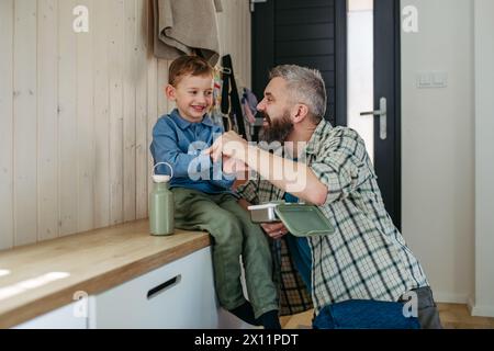 Father helping son get ready for kindergarten, preschool. Putting coat, shoes on, handing lunchbox with snacks. Stock Photo