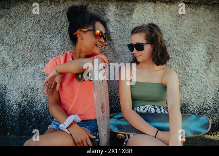 Teenager girl best friends with skateboards spending time outdoors in city during warm summer holiday day. Stock Photo