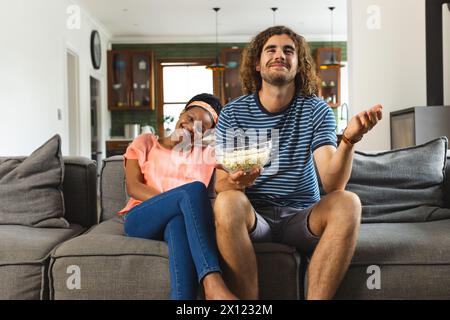 Diverse couple relaxing on couch, holding popcorn, watching television or movie at home Stock Photo