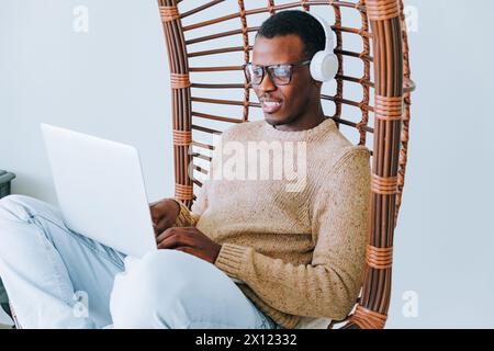 A contented African American man in a cozy sweater and headphones relaxes in a stylish wicker chair, engrossed in his laptop screen, exuding a vibe of comfortable productivity.  Stock Photo
