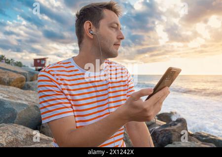Happy Caucasian young man listening to musc with headphones using smartphone on the sea coast and enjoying sunset. Handsome male outdoors using mobile Stock Photo