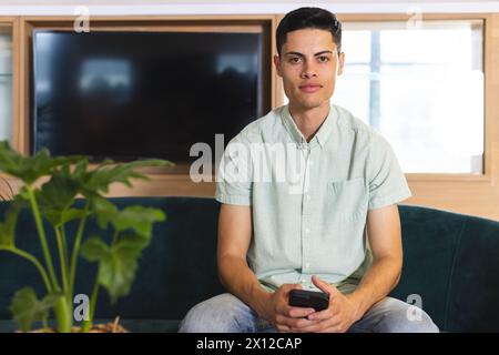 A young biracial man holding a smartphone sits on a couch in a modern business office Stock Photo
