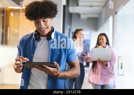 Biracial young man holding tablet in a modern business office, two women behind using tech Stock Photo