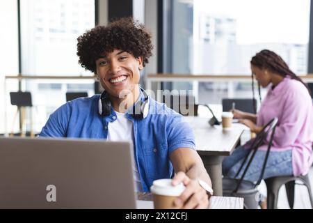 Biracial young man with headphones working on laptop in a modern business office, woman behind Stock Photo