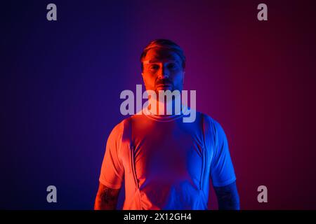 A man in smart glasses stands confidently in front of a vibrant purple and blue background in a futuristic studio setting. Stock Photo