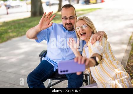 Man in wheelchair is spending time with his mother in park. They are taking selfie. Stock Photo