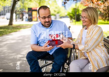 Man in wheelchair is spending time with his mother in park. He is receiving a present. Stock Photo