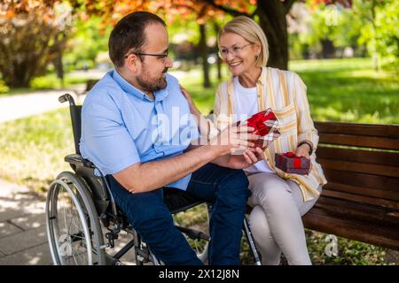 Man in wheelchair is spending time with his mother in park. He is receiving a present. Stock Photo