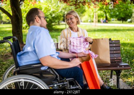 Man in wheelchair is spending time with his mother in park. They are talking after shopping. Stock Photo