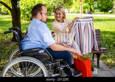 Man in wheelchair is spending time with his mother in park. They are talking after shopping. Stock Photo