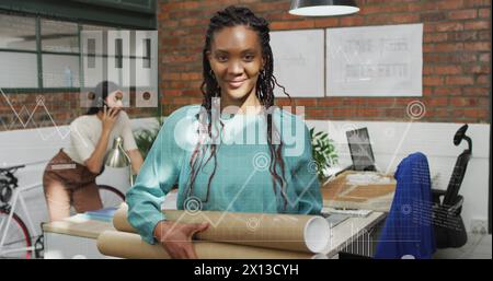 Image of graphs and icons over smiling biracial woman standing and holding cardboard rolls Stock Photo
