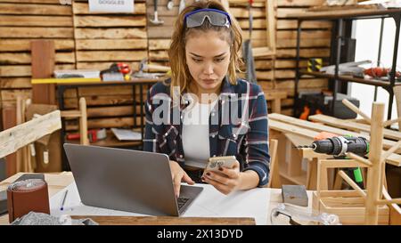 Young beautiful hispanic woman carpenter using laptop and smartphone at carpentry Stock Photo