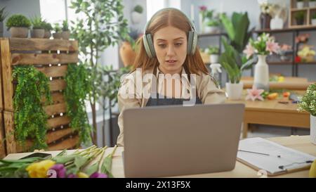 Caucasian redhead woman with headphones using laptop in florist shop surrounded by plants and flowers. Stock Photo