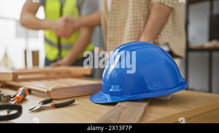 Two men shaking hands in a woodworking workshop with tools and a blue hardhat on the workbench Stock Photo