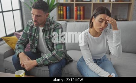 A worried hispanic couple sits apart on the sofa in a modern living room, reflecting tension and concern. Stock Photo