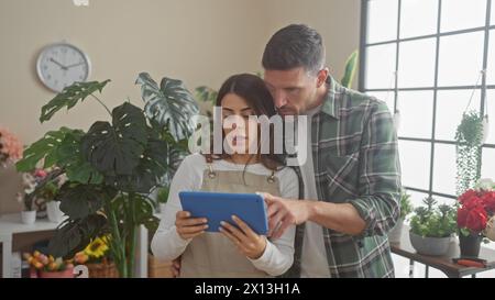 A man and woman, possibly florists, collaborate using a tablet amidst vibrant plants in an indoor flower shop. Stock Photo