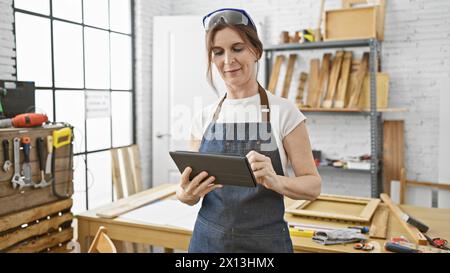 A middle-aged woman with short hair using a tablet in a bright, modern carpentry workshop. Stock Photo