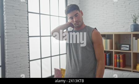 A tired hispanic man in a sleeveless shirt posing indoors near a window with a bookshelf and sofa in a modern apartment. Stock Photo