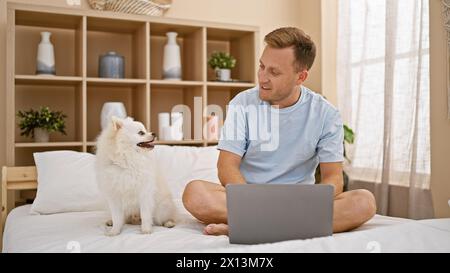 Chilled morning vibes with happy young caucasian man chilling on cozy bed in bedroom, relaxing with his pet dog and laptop. smiling, confident express Stock Photo