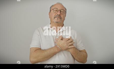 Serene middle-aged bald man with glasses and moustache in a white polo shirt, eyes closed, hands on chest, against a white background. Stock Photo
