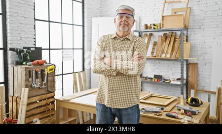 Confident middle-aged man with arms crossed stands in a well-equipped carpentry workshop, portraying craftsmanship and expertise. Stock Photo