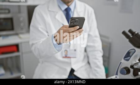 Handsome young hispanic scientist man, concentrated on work, analysing biology samples using smartphone technology in medical laboratory Stock Photo