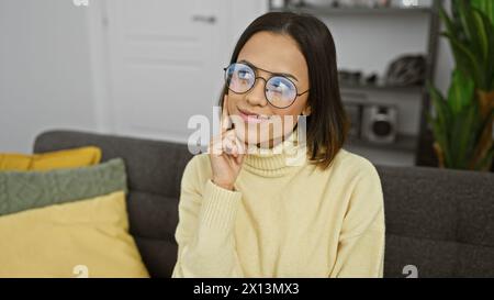 A content young hispanic woman with glasses, seated indoors, showcases a modern hairstyle and casual fashion at home. Stock Photo