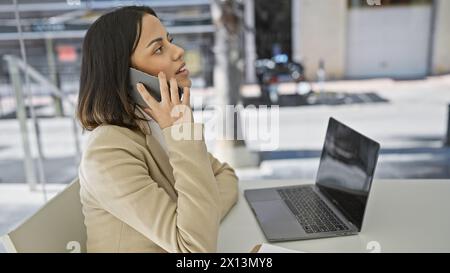 A young hispanic woman converses on a smartphone in a modern office setting, exemplifying professionalism against a blurred background. Stock Photo