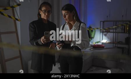 Two women investigators examine evidence using smartphones in the dimly lit room of a house crime scene. Stock Photo