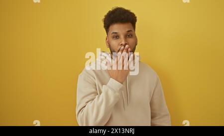 Adult black man covers mouth with hand against yellow background, portraying surprise, silence, or mistake. Stock Photo