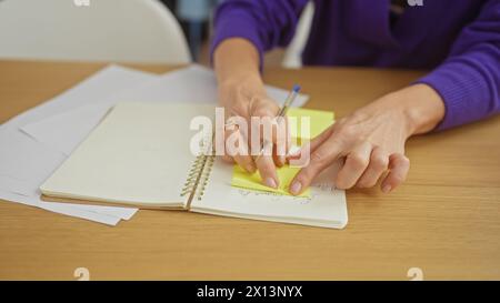 Caucasian woman writes on a sticky note at a wooden table, with paperwork and a notebook in a home setting. Stock Photo