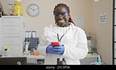 A smiling african american woman in lab coat and gloves using smartphone in a laboratory with equipment and a clock on the wall Stock Photo