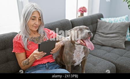 Relaxed grey-haired middle age woman, at home, sitting on comfortable sofa with pet dog, using touchpad in cozy living room Stock Photo