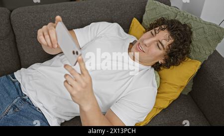 A young hispanic man with curly hair relaxes on a couch, smiling while using his phone in a cozy living room. Stock Photo