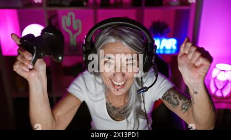 Confident grey-haired middle-aged woman streamer celebrating a big win while playing video game in her darkened gaming room at night Stock Photo