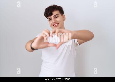 Young non binary man wearing casual white t shirt smiling in love doing heart symbol shape with hands. romantic concept. Stock Photo