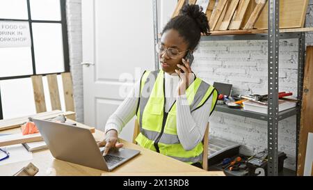 A focused african american woman multitasking in a carpentry studio, on the phone while using a laptop. Stock Photo