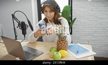 Young woman with headphones in a radio studio looking at phone by microphone and laptop, evoking a creative workspace. Stock Photo